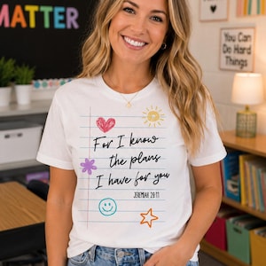 May include: A white short-sleeve t-shirt featuring a graphic of lined notebook paper with the text For I know the plans I have for you JEREMIAH 29:11. The design includes colorful hand-drawn doodles of a red heart, yellow sun, purple flower, blue smiley face, and orange star.