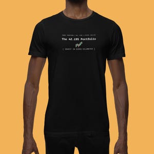 Op de afbeelding: Zwart T-shirt met witte tekst "The 42.195 Portfolio" boven een groen-witte afbeelding. Daaronder de zin "[ INVEST IN EVERY KILOMETER ]". Het shirt heeft ook de tekst "INDEX: MARATHON | VOL: KISH | STATUS: BULLISH".