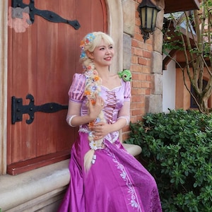 Könnte beinhalten: Eine Frau in einem lila-rosa Prinzessinnenkostüm mit einem langen blonden Zopf, der mit Blumen geschmückt ist. Das Kleid hat ein geschnürtes Oberteil und einen langen Rock. Ein grünes Frosch-Plüschtier sitzt auf ihrer Schulter.