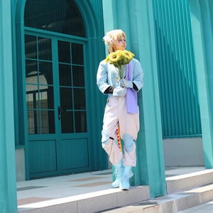 May include: A person in a light blue jacket, white pants, and blue boots holding sunflowers. The outfit has patches of blue and orange. The background features a teal door and wall.