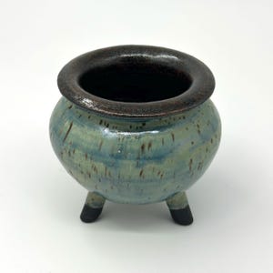 May include: A small, round ceramic pot with three short legs. The body features a mottled blue and green glaze with brown speckles, while the wide, thick rim is finished in a dark, textured brown glaze. The interior is dark and hollow.