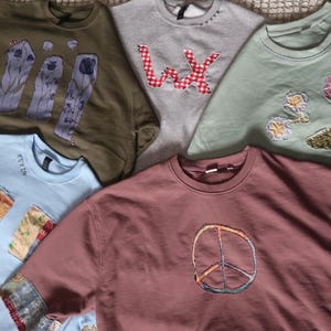 May include: An assortment of crew neck and long-sleeve shirts in various colors. Designs include a peace sign, floral embroidery, and patchwork details. The shirts are in shades of olive green, gray, light blue, and mauve.