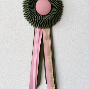 May include: A circular award rosette featuring a pleated olive green ribbon border and a central pink fabric-covered button. Three long ribbon tails hang from the bottom, including two solid pink ribbons, one olive green ribbon, and one gold glitter ribbon.