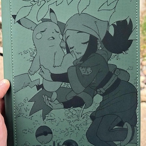 May include: A sage green rectangular card binder featuring a dark green line art illustration of a character hugging a Pikachu. The design includes foliage, two Poké Balls, and the text vaultx printed in the bottom left corner of the cover.