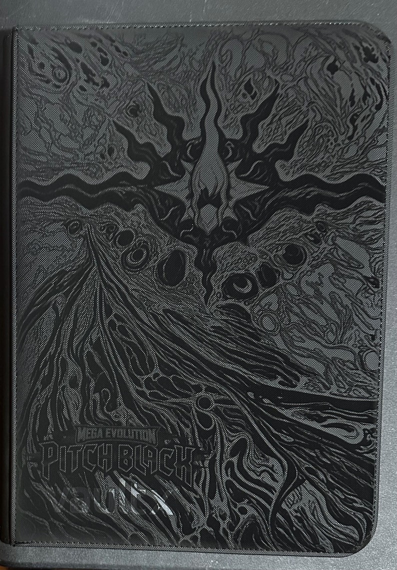 May include: A black rectangular binder featuring a dark, intricate, abstract graphic design across the front cover. The text "MEGA EVOLUTION" and "PITCH BLACK" is printed in a stylized font near the bottom, with a faint logo visible below the text.