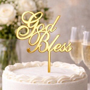 May include: A white cake with a gold "God Bless" cake topper. The cake is on a white cake stand, surrounded by flowers, candles, and champagne glasses. The cake has white frosting and a decorative border.