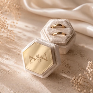 May include: A hexagonal beige velvet ring box featuring a gold-toned acrylic lid engraved with the letters A&V. The box is open, revealing two gold bands resting in the velvet slots. The lid is detached and positioned in front of the box.