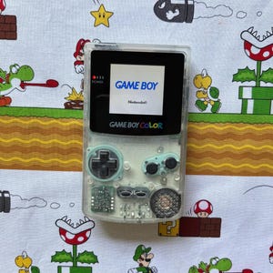 Op de afbeelding: Een transparante draagbare spelcomputer met een doorzichtige plastic behuizing, voorzien van een zwart scherm met de tekst GAME BOY en Nintendo. Het apparaat bevat een zwarte richtingsknop, twee lichtgroene knoppen met A en B, en een luidspreker.