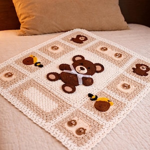 May include: A handmade, square baby blanket in cream, brown, and tan. The blanket features a central brown teddy bear applique with a white ribbon, surrounded by smaller bear and bee appliques. The blanket is placed on a bed.