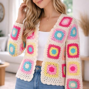 Può includere: Cardigan all'uncinetto color crema con motivi quadrati colorati. Ogni quadrato presenta un cerchio centrale in rosa, blu e giallo. Il cardigan ha un bordo smerlato e maniche lunghe. La modella indossa jeans blu e un top bianco.