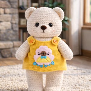 Op de afbeelding: Een handgemaakte, crèmekleurige gehaakte teddybeer met een gele jurk met een bloemenapplicatie. De beer heeft zwarte ogen, een bruine neus en oranje knoopaccenten. Het speelgoed staat op een beige tapijt.