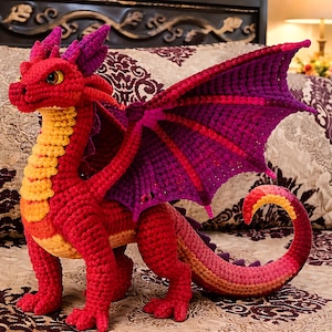 May include: A handmade crochet dragon in shades of red, orange, and purple. The dragon has large wings, a long tail, and yellow eyes. The dragon is sitting on a patterned bedspread.