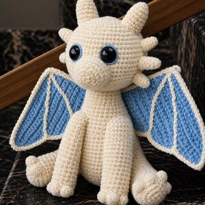 May include: A crocheted dragon plush toy in cream with blue wings and large black eyes. The handmade dragon has a friendly expression and is sitting on a dark surface. The wings are outlined in cream.