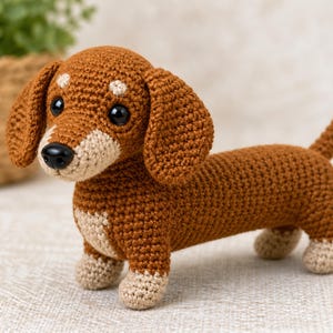 May include: A crocheted dachshund dog in shades of brown and beige. The handmade plush toy has a long body, floppy ears, and small legs. The dog has black eyes and a black nose. The toy is standing on a neutral surface.