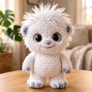 May include: A handmade, white crochet stuffed animal with a friendly expression. It has large brown eyes, blue accents on the ears, nose, and feet, and a fluffy white head. The plush toy is standing on a wooden surface.