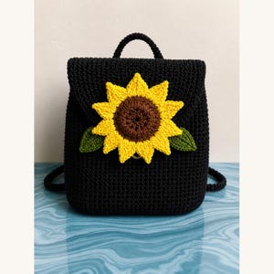 Könnte beinhalten: Schwarzer Häkelrucksack mit Sonnenblumenmuster. Die Sonnenblume hat gelbe Blütenblätter, eine braune Mitte und grüne Blätter. Der Rucksack hat einen oberen Griff und verstellbare Träger.
