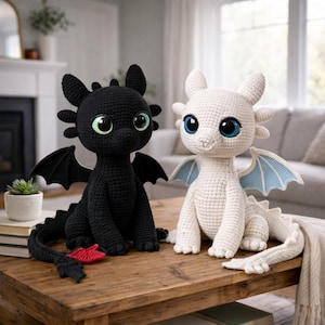 May include: Two crocheted dragon figures sit on a wooden table. One is black with green eyes, the other is white with blue eyes and light blue wings. Both have large, round eyes and detailed features, showcasing a handmade craft.