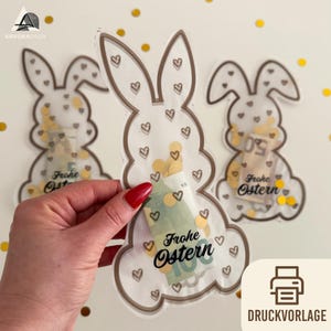 Può includere: Sacchetti regalo trasparenti a forma di coniglietto, con contorni marroni e dettagli a cuore, riempiti di denaro e coriandoli. Le borse hanno la scritta "Frohe Ostern" in nero. Coriandoli dorati punteggiano lo sfondo.