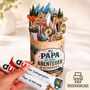 Könnte beinhalten: Eine Pop-up-Karte zum Vatertag mit Berg- und Camping-Motiv. Die Karte zeigt einen Kompass, eine Kamera und ein Schild mit der Aufschrift "Alles Gute zum Vatertag". Die Karte ist zylindrisch mit dem Text "Mit Papa ist jedes Abenteuer schöner".