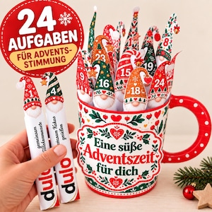 Może przedstawiać: Biały ceramiczny kubek w czerwono-zielone świąteczne wzory z napisem "Eine süße Adventszeit für dich", wypełniony znacznikami kalendarza adwentowego w kształcie krasnali ponumerowanymi od 1 do 24 oraz dwoma patyczkami "dulo".
