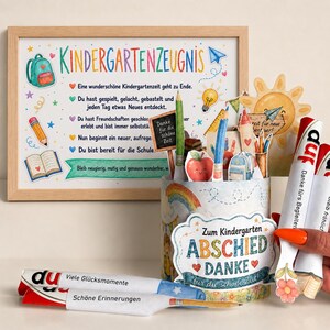 Può includere: Una stampa incorniciata intitolata Kindergartenzeugnis con illustrazioni colorate di materiale scolastico, accanto a un contenitore decorato pieno di rotoli di carta a forma di matita con testo in tedesco, tra cui Danke fürs Begleiten e Viele Glücksmomente.