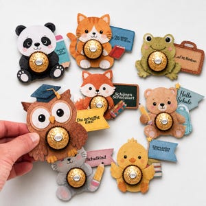 Puede incluir: Una colección de ocho figuras de papel troquelado con forma de panda, gato, rana, búho, zorro, oso, gatito y pollito, cada una sosteniendo un chocolate Ferrero Rocher dorado. Varias figuras incluyen textos y accesorios escolares.