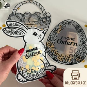 May include: A white Easter bunny-shaped decoration with the words "Frohe Ostern" and gold confetti inside. The bunny is held in front of a basket of Easter eggs and an egg-shaped decoration with the same text. The decorations are black and white.