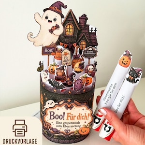 Könnte beinhalten: Ein zylindrischer Halloween-Behälter aus Papier mit einem Spukhaus, einem Geist und verschiedenen gruseligen Motiven. Das vordere Etikett trägt die Aufschrift "Boo! Für dich! Eine gespenstische süße Überraschung." Zwei weiße Röhrchen mit Halloween-Grafiken und deutschem Text werden daneben gehalten.