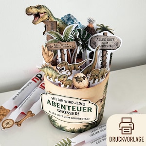 May include: A pop-up dinosaur-themed birthday card with various dinosaur figures, palm trees, and volcano illustrations. The card includes German text that reads "Alles Gute zum Geburtstag!" and "Mit dir wird jedes Abenteuer grösser!"