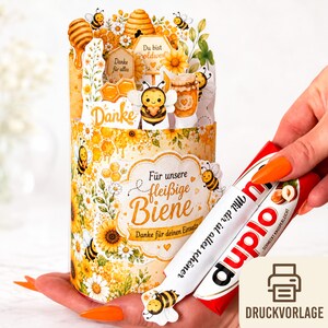Peut inclure: Un objet artisanal en papier jaune et blanc sur le thème des abeilles, avec des tournesols, des nids d'abeilles et des abeilles de dessin animé. Le mot "Danke" et d'autres textes allemands sont présents. Une barre chocolatée rouge et blanche est tenue à la main.