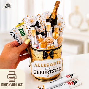 May include: A cylindrical gift box in gold and black with a bow, featuring cutouts of champagne, glasses, flowers, and a crown. The front label reads ALLES GUTE ZUM GEBURTSTAG. A hand holds a decorative paper insert with a 200 Euro banknote graphic.