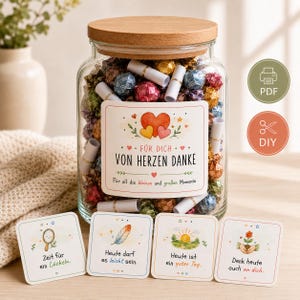 Könnte beinhalten: Ein klares Glasgefäß mit Holzdeckel, gefüllt mit bunten, in Folie eingewickelten Bonbons und kleinen Papierrollen. Das Etikett trägt die Aufschrift: FÜR DICH VON HERZEN DANKE. Vier quadratische Karten mit Illustrationen liegen vor dem Glas.