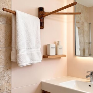 May include: A wooden towel rail with three rotating arms is mounted on a light pink wall. A white towel hangs from one arm. A small wooden shelf holds two white bottles. The bathroom features a white sink and a mirror.