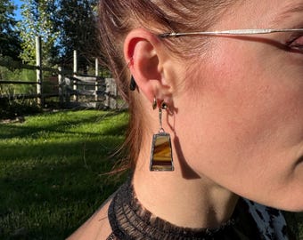 Könnte beinhalten: Ein trapezförmiger Ohrring aus Buntglas mit horizontalen Streifen in Braun-, Beige- und Cremetönen, eingefasst in einen dunklen Metallrahmen. Der Ohrring hängt an einem kleinen silberfarbenen Metallring, der an einem dünnen Ohrhaken befestigt ist.