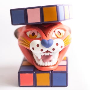 Puede incluir: Una cabeza de gato roja y caricaturesca con la boca muy abierta y dientes afilados, encima de una estructura de bloques coloridos. El gato tiene una nariz azul, manchas blancas y ojos grandes y amarillos. Una estructura de bloques similar está en su cabeza.