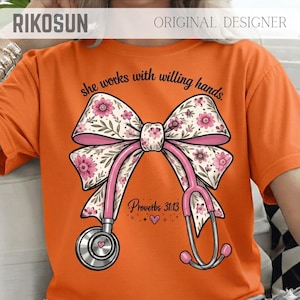 Könnte beinhalten: Orangefarbenes T-Shirt mit einem rosa Stethoskop in Form einer Schleife mit floralen Akzenten. Der Text "she works with willing hands" und "Proverbs 31:13" sind ebenfalls abgebildet. Ein Herzsymbol ist ebenfalls enthalten.