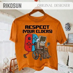 Puede incluir: Camiseta naranja con el texto "RESPECT (YOUR ELDERS)". El gráfico muestra un mando de videojuegos rojo y azul empujando una silla de ruedas con un personaje de mando de videojuegos. La camiseta tiene mangas cortas.