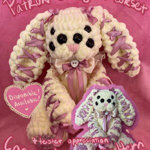 May include: A handmade, cream-colored crochet bunny with black spots, pink accents, and a pink bow. The bunny has long, floppy ears adorned with pink ribbons. The image includes the text "Patrón conejita corset" and "Corset bunny pattern". A smaller version of the bunny is also present.