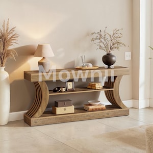 May include: A wooden console table with a curved design and two shelves. The table is a light brown color and has decorative items on top, including a lamp, vase, and framed pictures. The word "Mowm" is printed on the table.