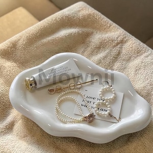Op de afbeelding: Een wit, wolk-vormig keramisch dienblad bevat een tube handcrème, een gouden haarclip, een parelketting, een paar parel oorbellen en een kaart met tekst. De lade staat op een beige oppervlak.