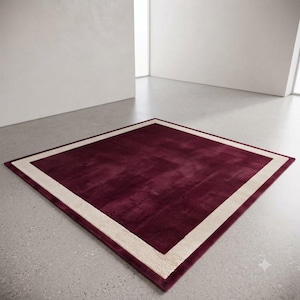 Könnte beinhalten: Ein quadratischer Teppich mit einem tief burgunderroten Mittelfeld, das von einem breiten, hellbeigen Rand umgeben ist. Der Teppich hat einen kurzen, dichten Flor und liegt flach, was ein klares, geometrisches Rahmendesign mit scharfen Ecken zeigt.