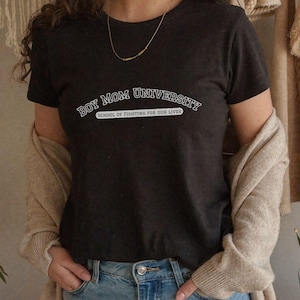 May include: A dark gray t-shirt with the text "BOY MOM UNIVERSITY" in a white, arched font. Below the main text is the phrase "SCHOOL OF FIGHTING FOR OUR LIVES" in a smaller, white font. The shirt is short-sleeved.