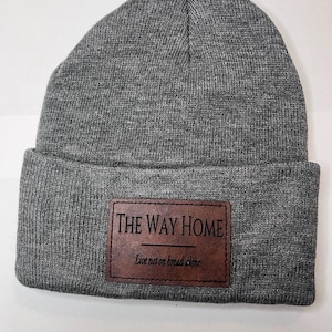 Könnte beinhalten: Graue Strickmütze mit einem braunen Lederaufnäher. Der Aufnäher zeigt den Text "THE WAY HOME" über der Linie und "Live not on bread alone" darunter. Ein klassischer, bequemer Stil.