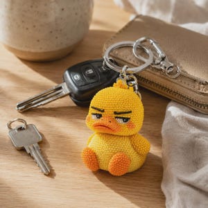 May include: A yellow duck keychain featuring a textured, beaded surface with a grumpy facial expression, including black eyebrows, eyes, and an orange beak. The duck is attached to a silver-toned metal split ring and a lobster claw clasp.