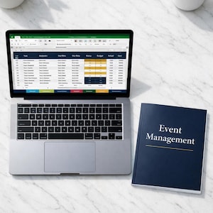 May include: A laptop displaying a spreadsheet sits next to a navy blue book titled "Event Management." A cup of coffee and a small potted plant are also on the white marble surface. The laptop is open, showing data in a spreadsheet format.