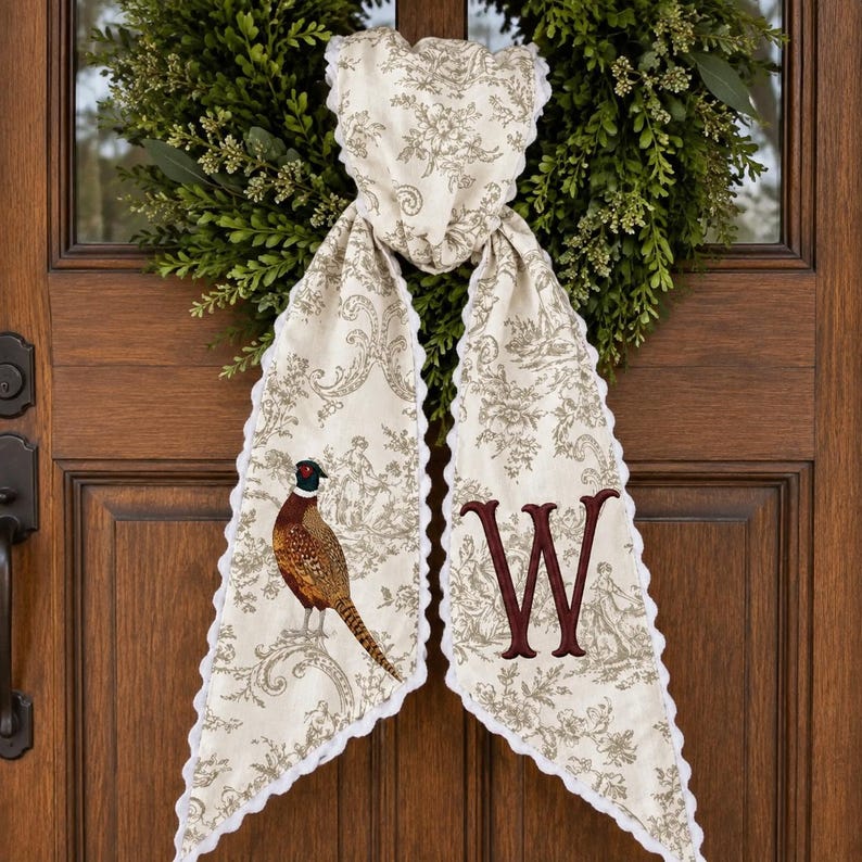 May include: A cream-coloured fabric door hanger featuring a brown toile pattern, a scalloped white edge, and a pheasant illustration on the left side. The right side displays a large, embroidered burgundy letter W in a serif font style.