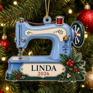 Könnte beinhalten: Ein hellblaues Nähmaschinen-Ornament mit einem cremefarbenen Etikett, auf dem der Name LINDA und das Jahr 2026 in dunkler Schrift stehen. Das Design umfasst eine rote Garnrolle, ein grünes Handrad und dekorative Stechpalmenblätter mit roten Beeren.