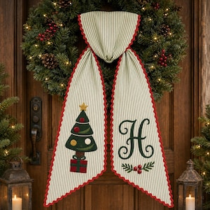 Peut inclure: Une suspension de porte en tissu rayé crème et vert avec un bord festonné rouge. Un côté présente un sapin de Noël brodé en vert avec une étoile dorée et un cadeau rouge. L'autre côté affiche une grande lettre H verte au-dessus de baies rouges.