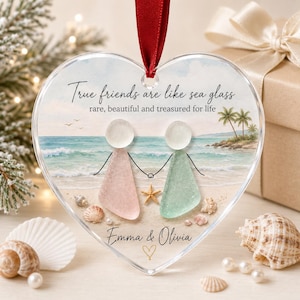 May include: A heart-shaped clear ornament featuring a beach scene with two figures made of sea glass, one pink and one light green. The text reads: True friends are like sea glass, rare, beautiful and treasured for life, Emma & Olivia.