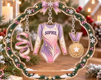 Può includere: Un ornamento circolare in acrilico trasparente con un bordo a ghirlanda festiva con fiocchi di neve e bacche. All'interno pendono un body a maniche lunghe con il nome SOPHIE, un nastro da ginnastica e una medaglia d'oro.